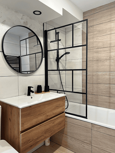 Baño con mueble de madera, espejo redondo negro y mampara de perfilería negra