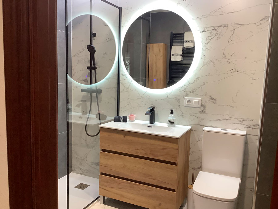 Después: Baño con espejo redondo iluminado y mueble de madera. Antes: baño antiguo con lavabo de pie y bañera.