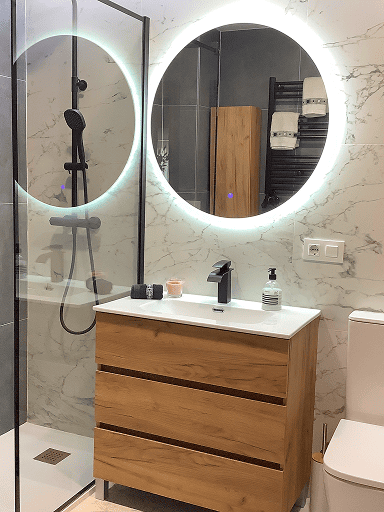 Baño con espejos redondos iluminados, mueble de madera y paredes de efecto mármol