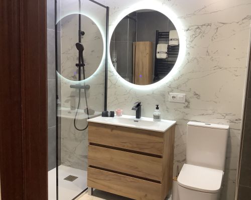 Después de la reforma: baño con mueble de madera, espejo redondo iluminado y paredes de efecto mármol